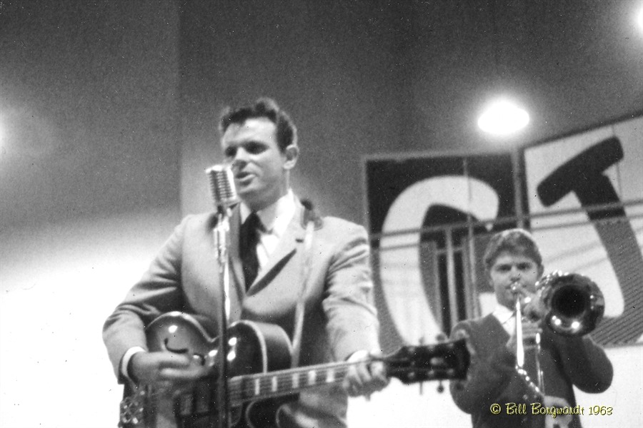 Black and White photo of 2 men playing 1960's pop music. The man in the foreground is playing a guitar and singing, the man in the background is playing trombone.