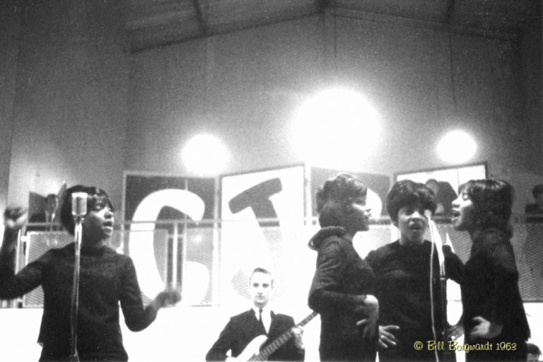 Black and White image of 1963 girl group. On the left is one girl dancing and singing, in the middle is a man playing guitar and on the right are three women singing.