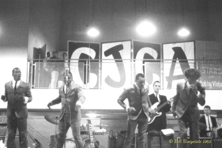 Black and White image of a 1963 rock band playing and dancing to the music. CJCA is in large letters in the background