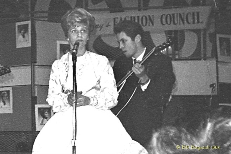 Black and white photo, 1963, woman in white dress stands at the microphone.