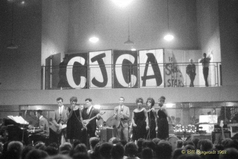 Black and White photo. Large band performs, with 3 back up singers and 3 musicians. A crowd listens.