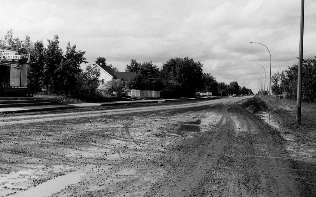 B&W photo of 149th Street looking north from 97th Avenue in 1960; road is gravel; sign for a grocery store is on the left.